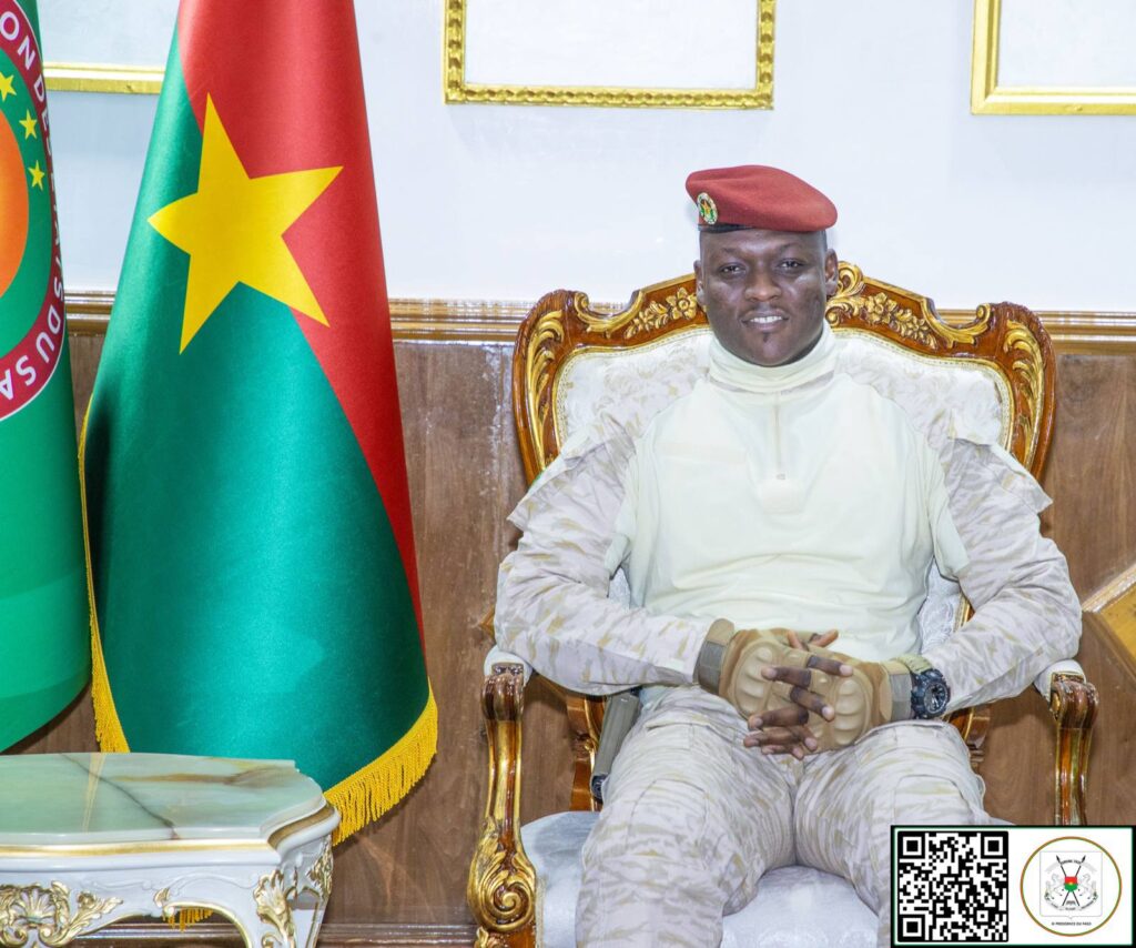 Le président Ibrahim Traoré assis sur un fauteuil, souriant, devant les drapeaux du Burkina Faso et de l’Union des États du Sahel.