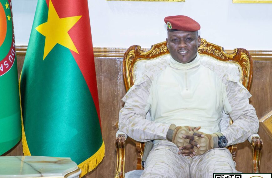 Le président Ibrahim Traoré assis sur un fauteuil, souriant, devant les drapeaux du Burkina Faso et de l’Union des États du Sahel.