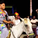 Une artiste haïtienne défile à cheval lors d’un événement nocturne, vêtue d’une tenue colorée et entourée de spectateurs