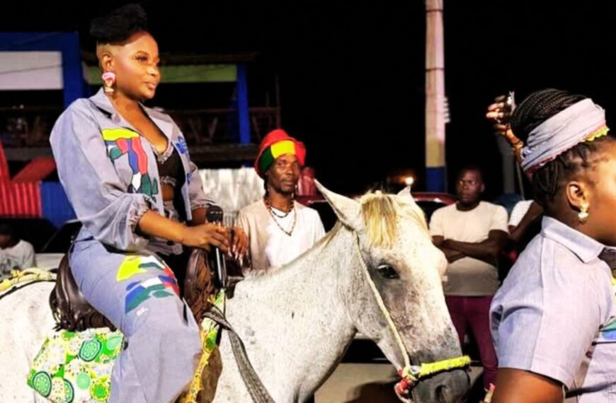 Une artiste haïtienne défile à cheval lors d’un événement nocturne, vêtue d’une tenue colorée et entourée de spectateurs