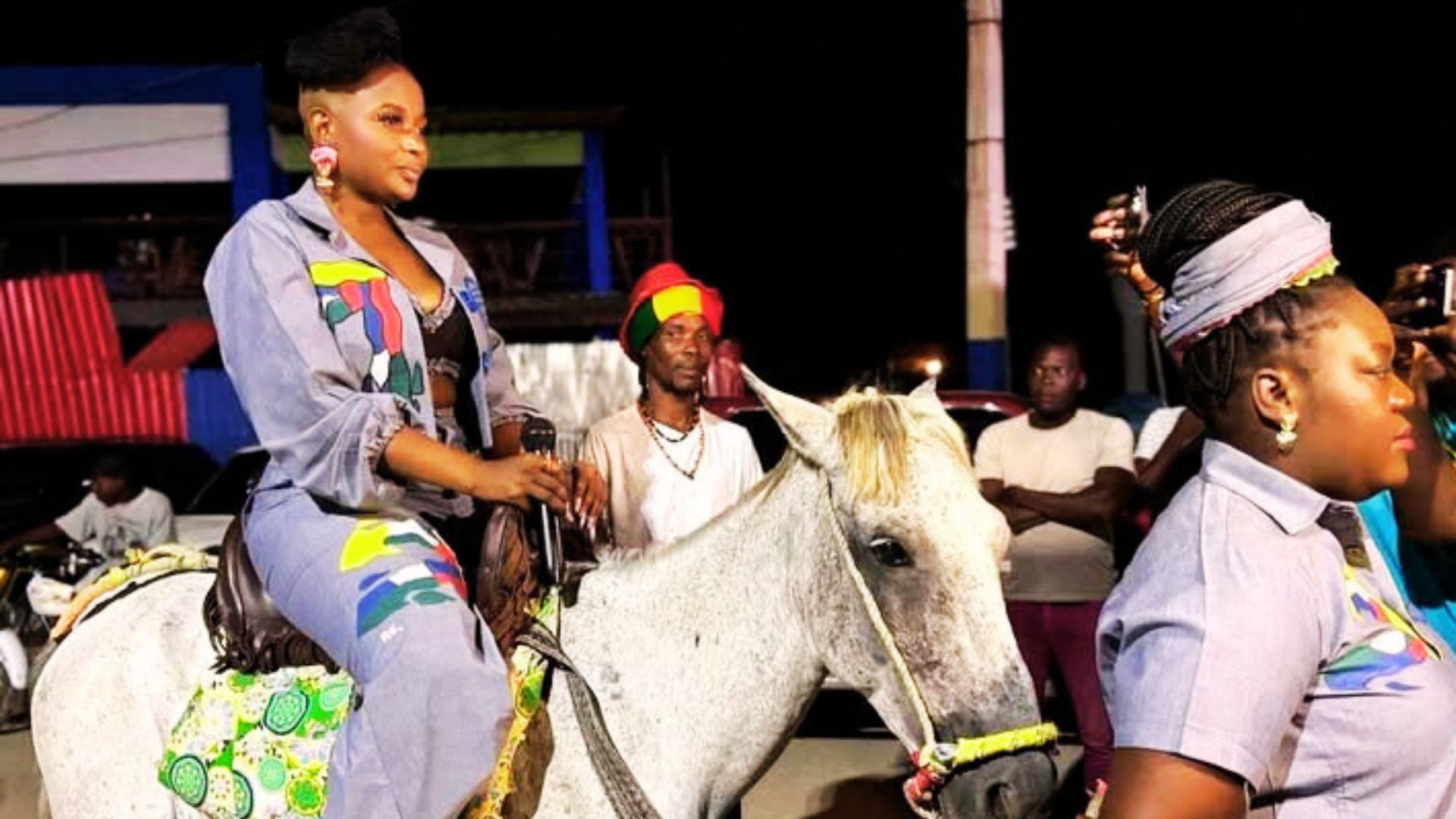 Une artiste haïtienne défile à cheval lors d’un événement nocturne, vêtue d’une tenue colorée et entourée de spectateurs
