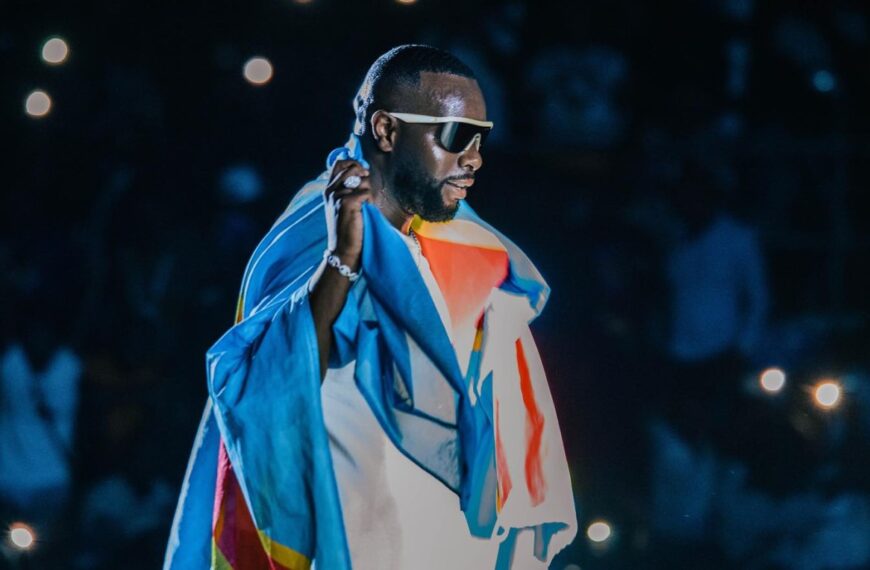 Gims drapé du drapeau congolais lors d’un concert, sous les lumières du public.