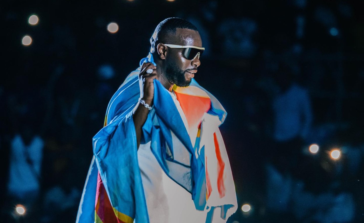 Gims drapé du drapeau congolais lors d’un concert, sous les lumières du public.