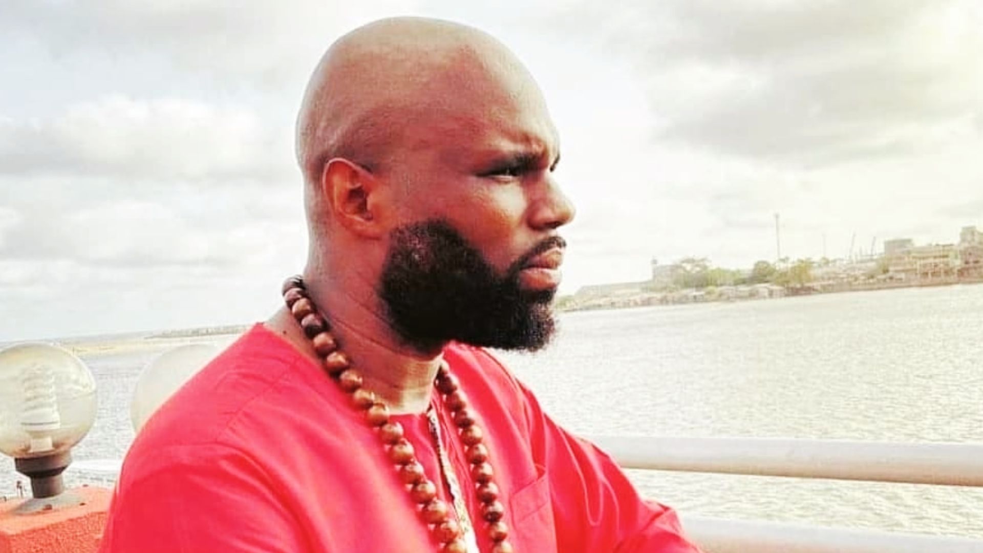 Kemi Seba regarde l’horizon, vêtu d’une tunique rouge, sur un pont en bord de mer.