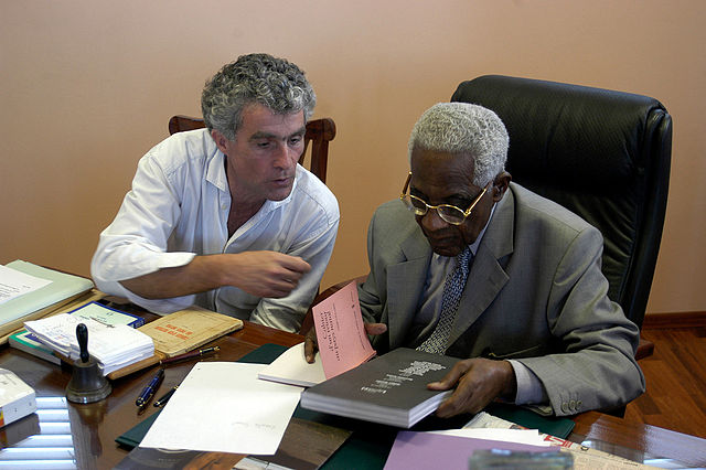 Deux hommes assis à un bureau, échangeant autour de livres, dont l’un est Aimé Césaire, figure majeure du mouvement de la Négritude.