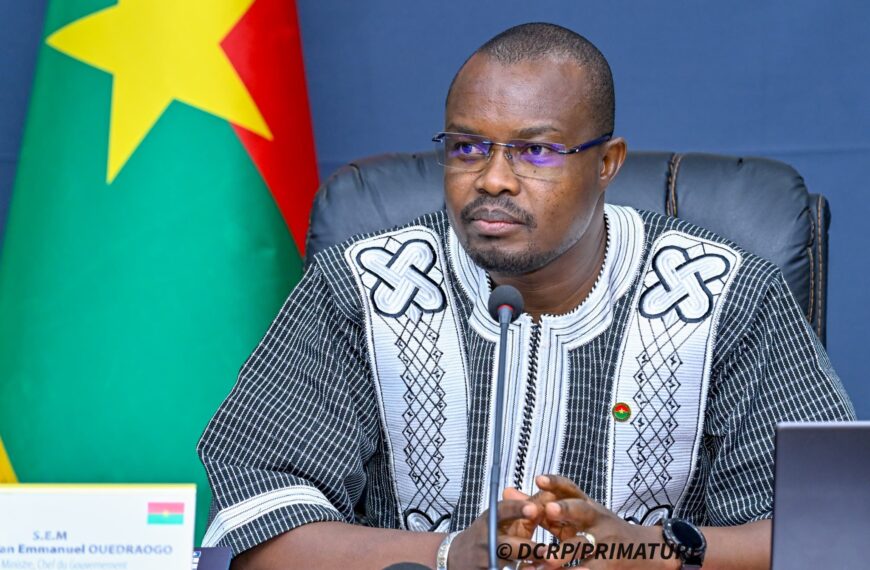 Portrait du Premier ministre du Burkina Faso, Rimtalba Jean Emmanuel Ouédraogo, s’exprimant lors d’une réunion officielle à Ouagadougou, avec le drapeau national en arrière-plan.