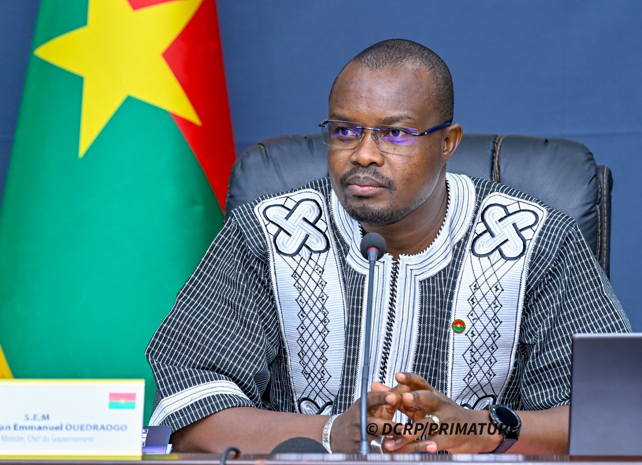 Portrait du Premier ministre du Burkina Faso, Rimtalba Jean Emmanuel Ouédraogo, s’exprimant lors d’une réunion officielle à Ouagadougou, avec le drapeau national en arrière-plan.