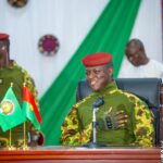 Le capitaine Ibrahim Traoré assis derrière un bureau, en tenue militaire, devant des drapeaux posés sur la table lors d’une rencontre officielle.
